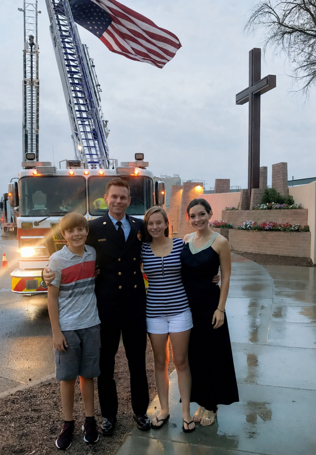 Scott Robb in fire-service dress uniform with his family, a fire engine, raised ladders, an American flag, and a cross